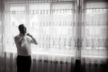 Bir adam pencerenin önünde giyiniyor. Perdeler beyaz ve pencere içeri biraz ışık giriyor. Adam beyaz gömlek ve siyah pantolon giyiyor.