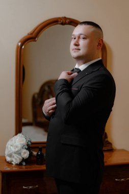 Takım elbiseli bir adam aynanın önünde kravatını düzeltiyor. Formalite ve detaylara dikkat etme kavramı, adamın görünüşünün parlak ve profesyonel olması için zaman ayırması gibi.