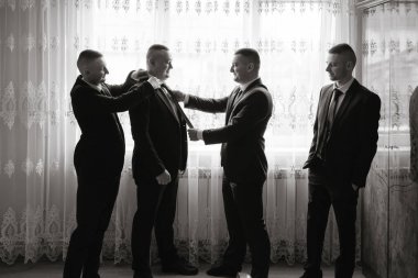 Takım elbiseli dört adam pencerenin önünde dikiliyor ve bir adam diğerinin kravatını düzeltiyor. Erkekler takım elbise ve kravat giydiği için sahne resmi ve profesyonel. Görüntünün ayarları