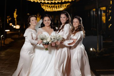 Beyaz elbiseli dört kadın fotoğraf için poz veriyor. Kadınlardan biri elinde bir buket çiçek tutuyor.