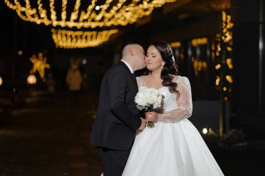 Bir gelin ve damat ışıklı bir tabelanın önünde öpüşüyorlar. Gelin elinde bir buket çiçek tutuyor.