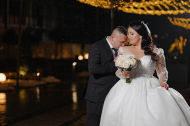 Bir gelin ve damat yağmurda öpüşüyor. Gelin elinde bir buket çiçek tutuyor.