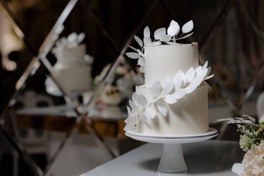 Üzerinde yaprak deseni olan beyaz bir pasta beyaz bir tabakta oturur. Pasta bir masaya konur ve bir aynayla çevrilidir.