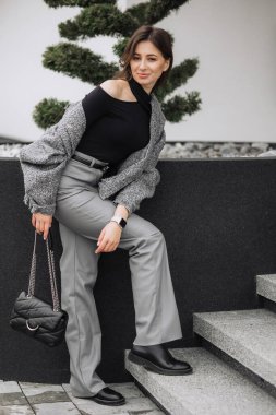 Siyah ve gri elbiseli bir kadın fotoğraf için poz veriyor. O, siyah bir ceket ve gri pantolon giyiyor. O, siyah bir el çantası tutuyor ve bir duvara yaslanıyor. Görüntü sıradan.