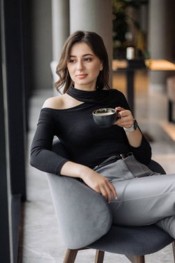 Bir kadın elinde bir fincan kahveyle bir sandalyede oturuyor. O, siyah bir gömlek ve gri pantolon giyiyor. Rahatlama ve rahatlama kavramı