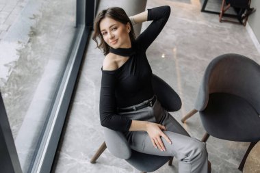 Kolları çapraz bir şekilde bir sandalyede oturan ve yüzünde bir gülümseme olan bir kadın. Siyah bir bluz ve gri bir pantolon giyiyor.