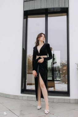 Siyah elbiseli bir kadın pencerenin önünde duruyor. Yüksek topuklu ayakkabı giyiyor ve elleri kalçasında. Elbise uzun ve ortadan ikiye bölünmüş. Sahne zarif ve sofistike.