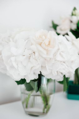 Masanın üzerinde bir vazo beyaz çiçek duruyor. Çiçekler bembeyaz ve onları çiçek açmış gibi gösterecek şekilde düzenlenmiş. Vazo temiz ve çiçeklerin içinden görünmesini sağlıyor.