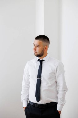 Beyaz gömlekli ve siyah kravatlı bir adam beyaz bir duvarın önünde duruyor. Bir şey ya da birini bekliyor.