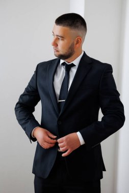 Takım elbiseli ve kravatlı bir adam beyaz bir duvarın önünde duruyor. Kravatını düzeltiyor ve kameraya bakıyor. Formalite ve profesyonellik kavramı. Çünkü adam takım elbise ve kravat giymiş.