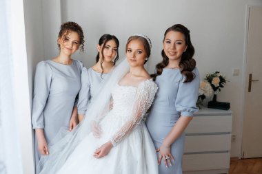 Dört kadın pencerenin önünde poz veriyor. Gelin beyaz bir elbise ve duvak takıyor. Diğer kadınlar mavi giyinmiş. Sahne mutlu ve kutlama dolu.