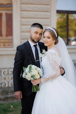 Bir gelin ve damat binanın önünde fotoğraf için poz veriyorlar. Damat takım elbise ve kravat takarken gelin beyaz bir elbise ve saç bandı takıyor. Bir buket tutuyorlar.