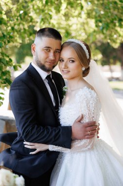 Bir gelin ve damat fotoğraf için poz veriyor. Gelin beyaz bir elbise ve duvak takıyor. Sahne mutlu ve kutlama dolu.