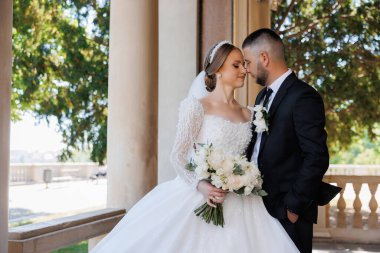 Gelin ve damat bir balkonda duruyorlar, gelin bir buket çiçek tutuyor. Sahne romantik ve mutlu, çünkü çift birbirini kucaklıyor ve gözlerinin içine bakıyor.