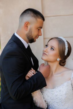 Bir adam ve bir kadın fotoğraf için poz veriyorlar, takım elbiseli bir adam ve gelinlik giyen bir kadın. Adam kadının elini tutuyor ve ikisinin de yüzünde bir gülümseme var.