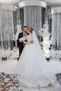 Bir gelin ve damat, şelaleli beyaz bir perdenin önünde duruyorlar. Gelin beyaz bir elbise giyiyor ve damat bir takım elbise giyiyor. El ele tutuşup kameraya gülümsüyorlar.