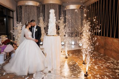 Bir gelin ve damat düğün pastasının önünde duruyorlar. Gelin beyaz bir elbise giyiyor ve damat bir takım elbise giyiyor. Sahne mutlu ve kutlama dolu.