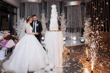 Bir gelin ve damat düğün pastasının önünde duruyorlar. Gelin uzun beyaz bir elbise giyiyor ve damat siyah bir takım elbise giyiyor. Pasta beyaz kremayla süslenmiş ve altın kaplamalı.