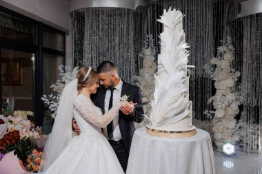 Bir gelin ve damat düğün pastasını birlikte kesiyorlar. Pasta beyaz ve üzerinde tüy tasarımı var. Çift çiçeklerle ve diğer dekorasyonlarla çevrili, neşeli bir atmosfer yaratıyor.