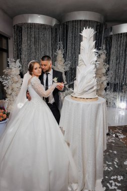 Bir gelin ve damat düğün pastasını birlikte kesiyorlar. Gelin beyaz bir elbise giyiyor ve damat bir takım elbise giyiyor. Pasta beyaz kremayla süslenmiş ve üstünde bir tasarım var.