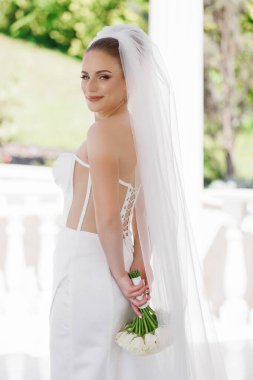 Bir gelin, bir buket beyaz çiçekle fotoğraf çektirmek için poz veriyor. Beyaz bir elbise ve duvak takıyor. Mutluluk ve kutlama kavramı, gelin gelinliğini giydiği gibi.