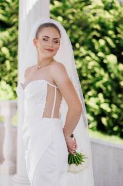 Beyaz elbiseli bir kadın elinde bir buket çiçek tutuyor. Gülümsüyor ve kameraya bakıyor. Mutluluk ve kutlama kavramı. Kadın büyük ihtimalle bir düğüne ya da özel bir etkinliğe katılıyor.