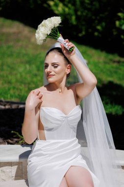 Beyaz bir elbise giyen ve elinde bir buket beyaz çiçek tutan bir kadın. Gülümsüyor ve kameraya bakıyor.