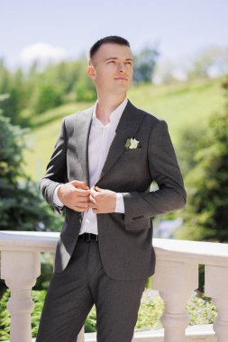 Takım elbiseli ve kravatlı bir adam balkonda dikilip manzaraya bakıyor. Beyaz gömlek ve siyah kuşak giyiyor.