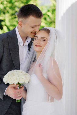 Bir gelin ve damat birbirlerine sarılıp öpüşüyorlar. Gelin beyaz bir elbise giyiyor ve damat da takım elbise giyiyor.