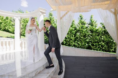 Bir gelin ve damat bir binanın merdivenlerinden çıkıyorlar. Gelin bir buket tutuyor ve damat elini tutuyor. Sahne romantik ve mutlu.