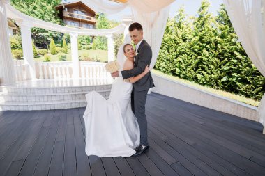 Gelin ve damat ahşap bir güvertede, gelin bir buket tutuyor. Sahne mutlu ve romantik, çünkü çift düğünlerini kutluyor.