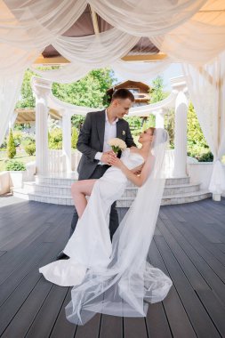 Gelin ve damat, çardağın önünde fotoğraf çektirmek için poz veriyorlar. Gelin bir buket tutuyor ve damat bir çiçek tutuyor. Sahne mutlu ve romantik.