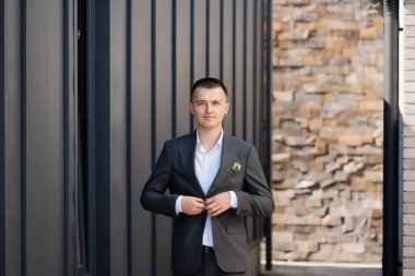 Takım elbiseli bir adam tuğla bir duvarın önünde duruyor. Yakasında yeşil bir rozet var.