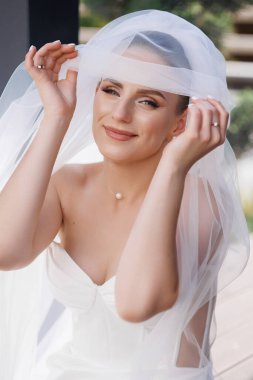 Bir kadın beyaz bir peçe takıyor ve gülümsüyor. İnci bir kolye takıyor ve saçları topuz şeklinde.