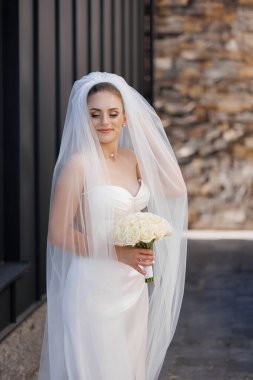 Başında peçesi ve elinde buketi olan bir gelin binanın önünde duruyor.