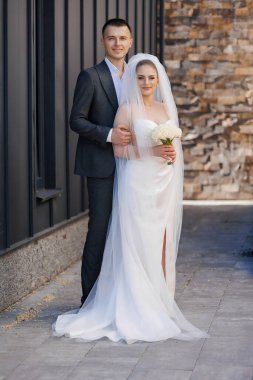 Bir gelin ve damat tuğla bir duvarın önünde fotoğraf için poz veriyorlar. Damat takım elbise giyerken gelin beyaz bir elbise ve peçe takıyor. Bir buket tutuyorlar.