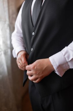 Siyah yelek ve beyaz gömlekli bir adam giyinmeye hazırlanıyor. Kravatını düzeltiyor ve bir yüzük takıyor. Özel bir durum için formalite ve hazırlık kavramı