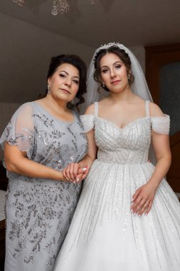 Gelinlikli bir kadın annesinin yanında duruyor. Anne gri bir elbise giyiyor ve kız beyaz bir elbise giyiyor. Sahne mutlu ve kutlama dolu.