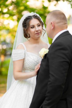 Gelin ve damat parkta dikiliyor, gelin beyaz bir elbise giyiyor ve damat siyah bir takım elbise giyiyor.