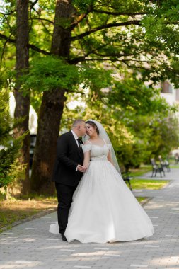 Bir gelin ve damat parkta bir patikada yürüyorlar. Gelin beyaz bir elbise giyiyor ve damat bir takım elbise giyiyor. El ele tutuşup kameraya gülümsüyorlar. Sahne mutlu ve romantik.