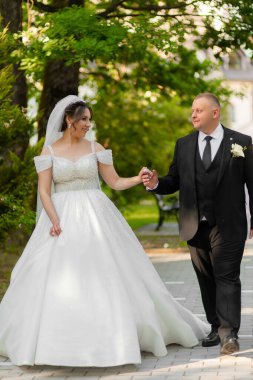 Bir gelin ve damat parkta bir patikada yürüyorlar. Gelin beyaz bir elbise giyiyor ve damat siyah bir takım giyiyor.