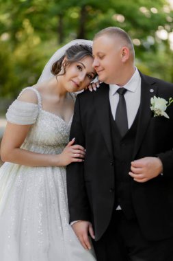 Bir gelin ve damat parkta birbirlerine sarılıyorlar. Gelin beyaz bir elbise giyiyor ve damat siyah bir takım giyiyor.