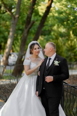 Gelin ve damat bir köprüde duruyorlar, gelin beyaz bir elbise ve damat siyah bir takım elbise giyiyor.