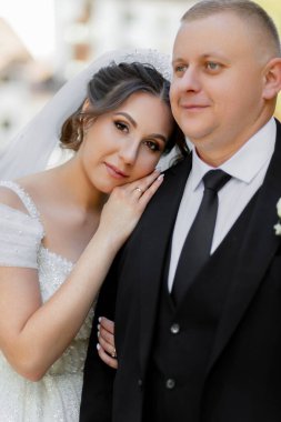 Bir gelin ve damat fotoğraf için poz veriyor. Gelin beyaz bir elbise giyiyor ve damat siyah bir takım elbise giyiyor. İkisi de gülümsüyor ve mutlu görünüyorlar.