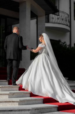 Gelin ve damat kırmızı halıda yürüyorlar. Gelin beyaz bir elbise ve duvak takıyor.