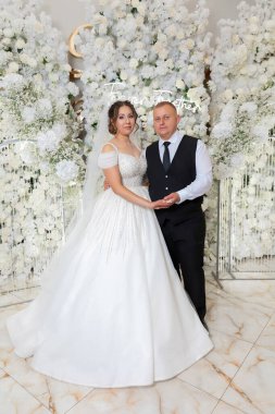 Bir gelin ve damat beyaz bir duvarın önünde poz veriyorlar. Gelin beyaz bir elbise giyiyor ve damat siyah bir yelek giyiyor. Sahne mutlu ve kutlama dolu.