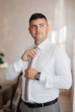 Beyaz gömlekli ve gri pantolonlu bir adam kravatını düzeltiyor. Saat takıyor ve yüzünde ciddi bir ifade var.