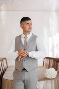 Takım elbiseli ve yelekli bir adam elinde bir vazoyla masanın önünde duruyor.