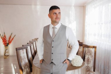 Takım elbiseli ve yelekli bir adam sandalyeli uzun bir masanın önünde duruyor. Kravat takıyor ve fotoğraf için poz veriyor. Odanın resmi ve zarif bir atmosferi var.