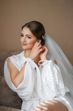 Bir kadın kanepede oturuyor ve saçını düzeltiyor. Beyaz bir elbise ve duvak takıyor.